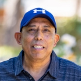 Man with dark skin wearing a blue hat leaning his left arm on a large rock with writing on it.